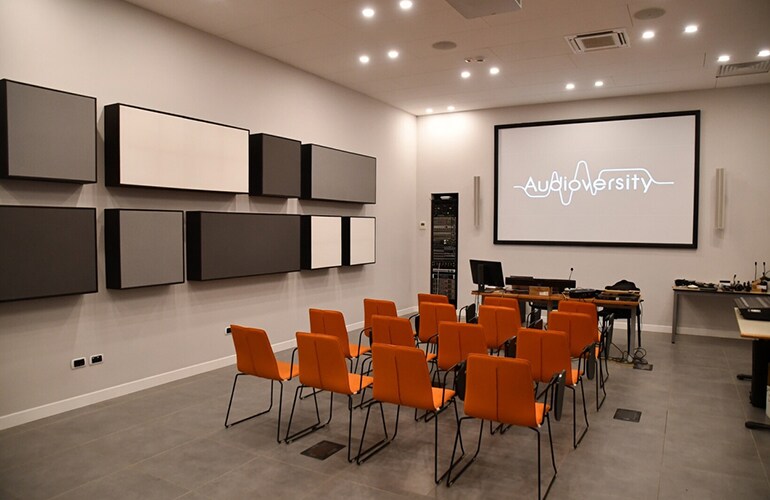 Centre de formation de Lesmo avec chaises orange, écran de présentation et équipement audio mural.